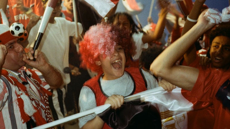 A group of enthusiastic fans cheer energetically, with one wearing a red and white wig. The scene is lively, filled with excitement and joy.
