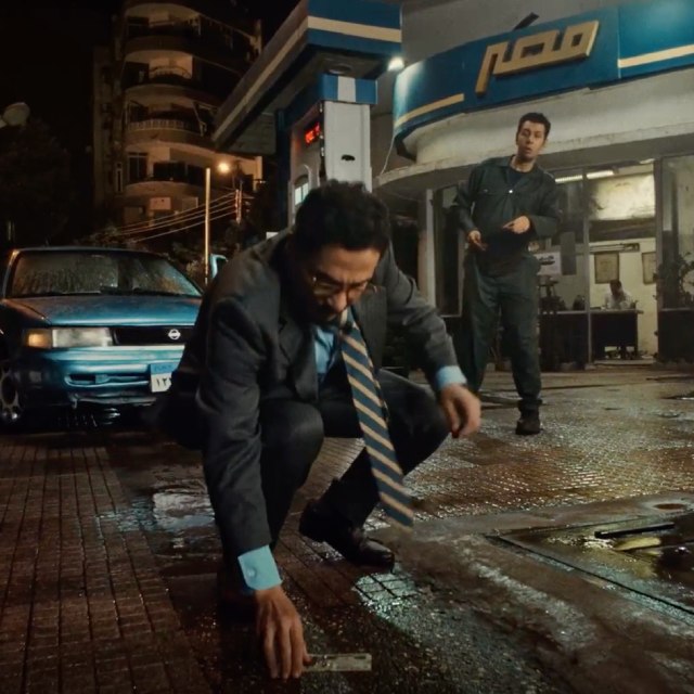 Man in a suit picking up a banknote from a wet sidewalk outside a bank at night