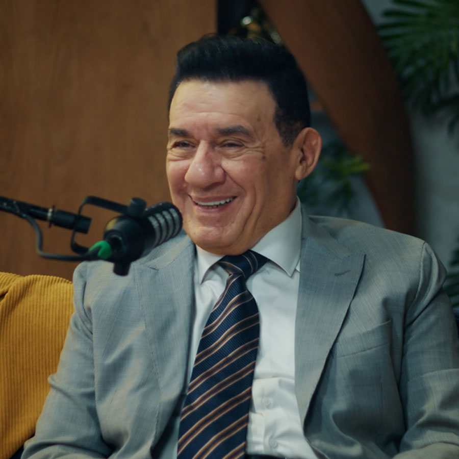 Medhat Shalaby in a gray suit and striped tie smiles while sitting on a blue couch. A microphone is in front of him, with plants and wood paneling in the background.