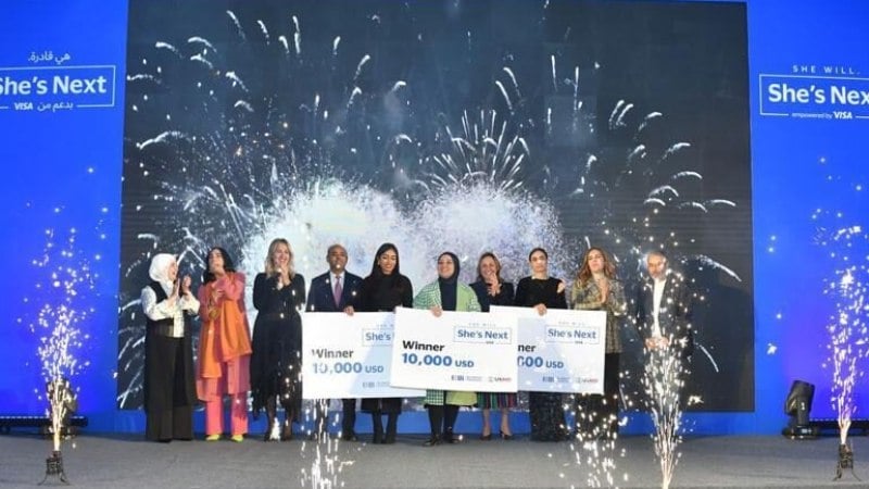 A group of people stand on stage holding large checks for $10,000, celebrating. Fireworks and a "She's Next" Visa banner add excitement to the scene.