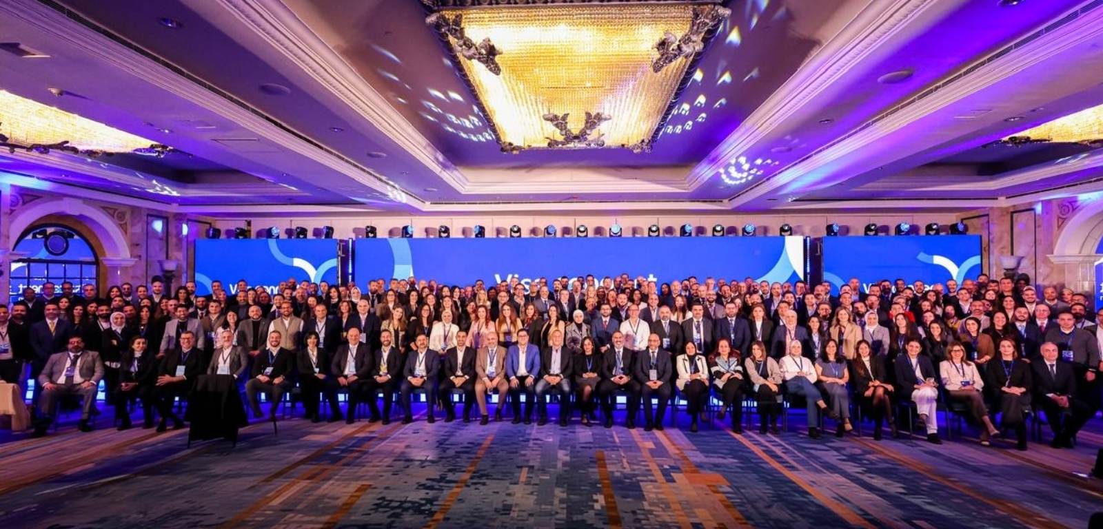 A large group of people gathered in a conference hall beneath ornate chandeliers with blue lighting.